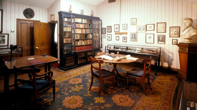 An image of the attic at Carlyle's House in London set out as it was when Carlyle wrote their using it as his study. It is a light room with wooden doors, large dark wood bookcase, tables and glass display case